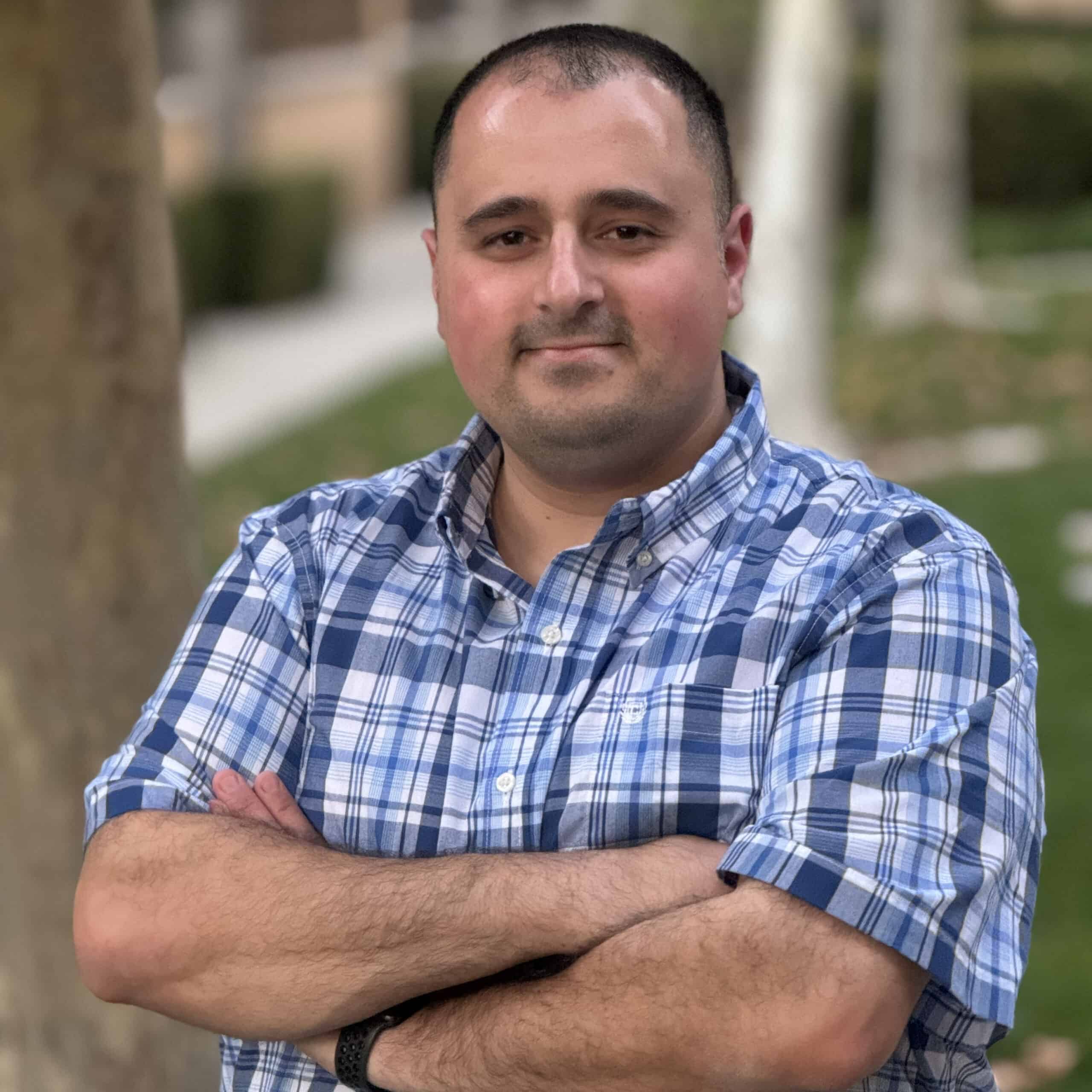 Bobby Omari wearing a plaid button up t-shirt smiling with crossed arms with a park behind him.